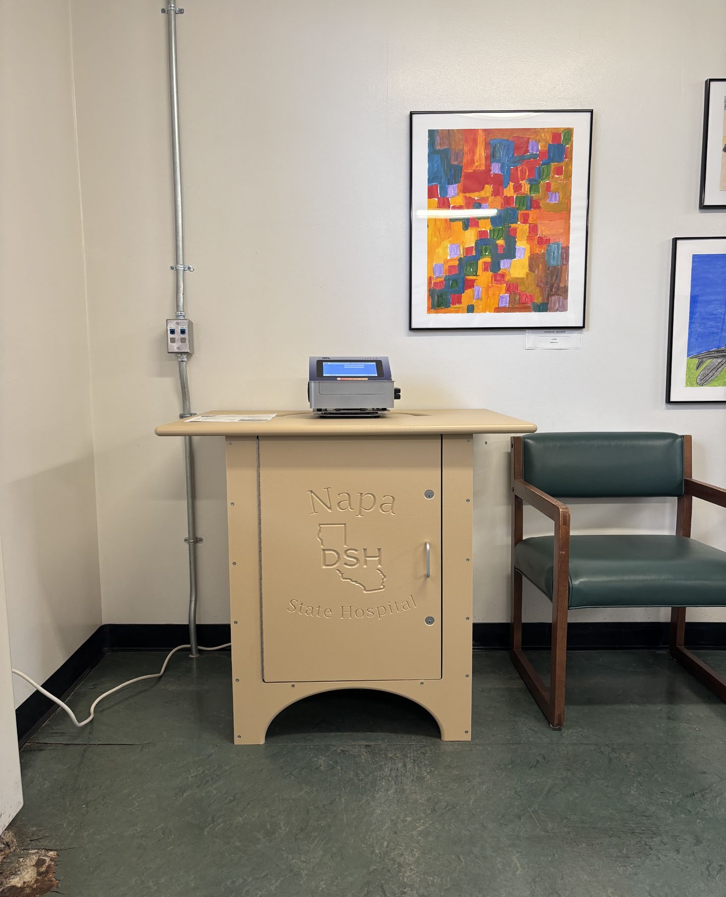 
			An image of one of the hospitals two credit card deposit terminals consisting of a card machine mounted on a tan cabinet engraved with the hospital’s logo, located in the Program 4 Visitor Center beside a green-upholstered chair with patient artwork on the wall behind it.